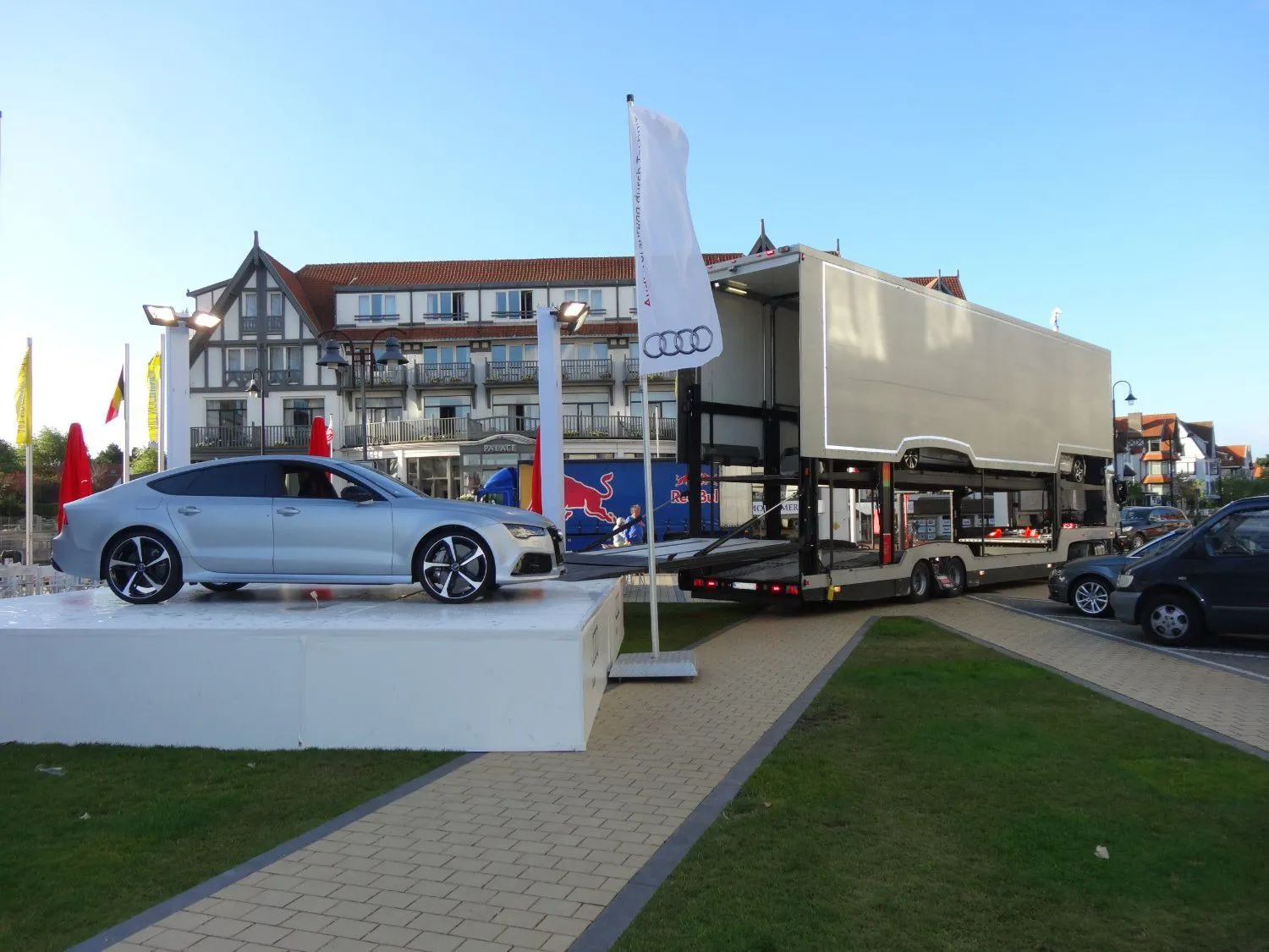 Zilverkleurige Audi Sportback op een witte sokkel bij een buitenevenement, transporter lost erachter