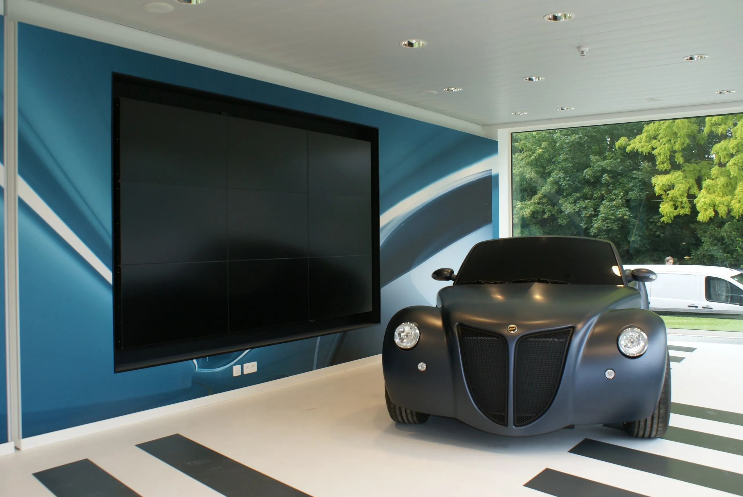 Donkere conceptcar op een sokkel in een showroom, groot scherm erachter