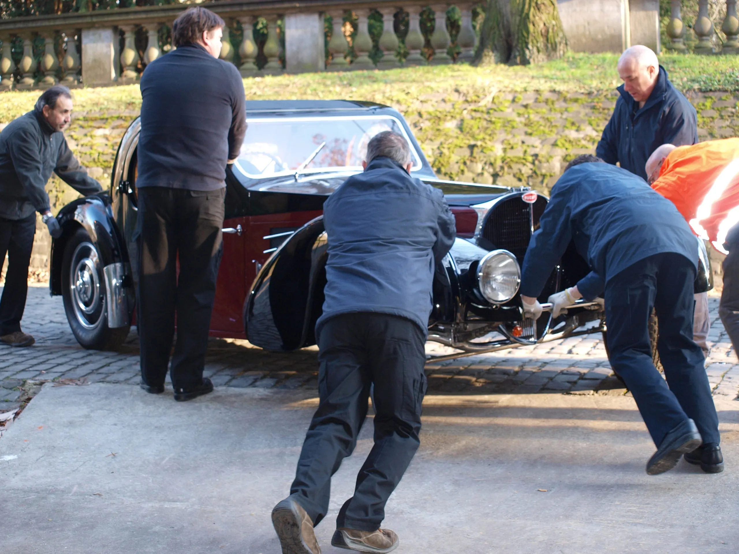 Vier mannen duwen een vooroorlogse Bugatti met de hand over de kasseien langs een park