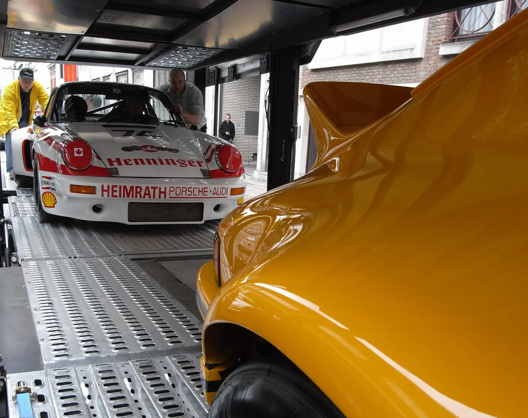 A race Porsche 911 pushed aboard an e-motion transporter by two crew members, the wing of a yellow 911 in the foreground