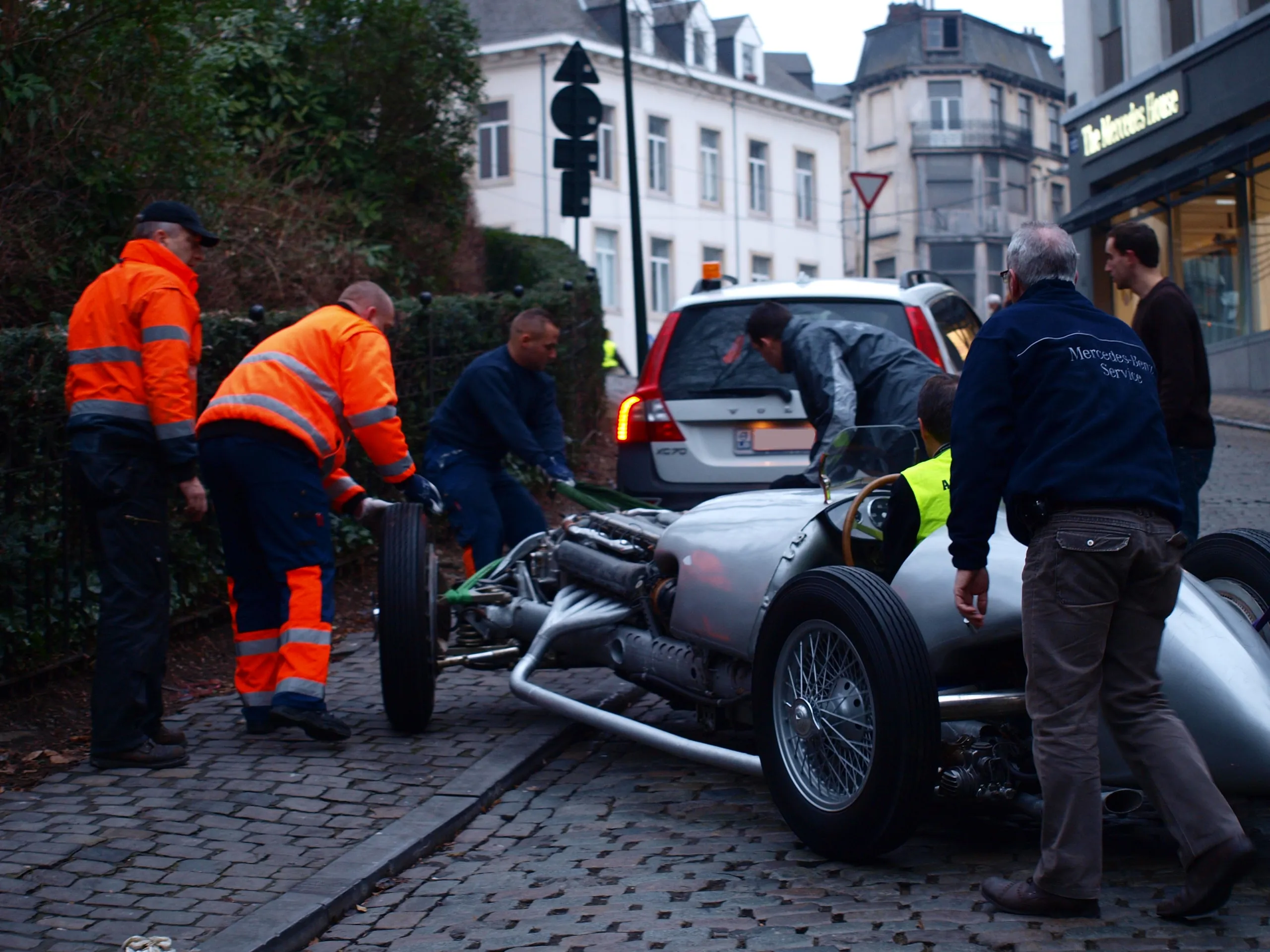 Ploeg in fluohesjes duwt een vooroorlogse Mercedes-grandprixwagen over de kasseien