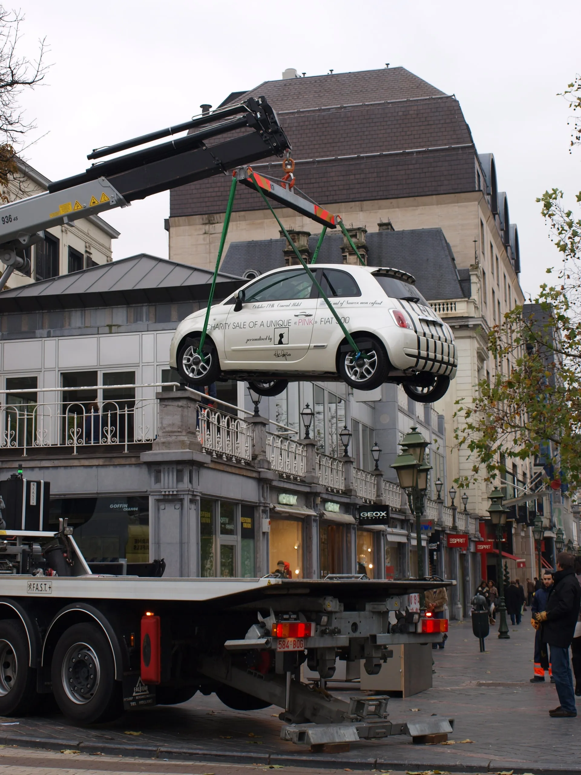 Fiat 500 hangend aan de kraanarm boven de laadbak, gebouwen eromheen