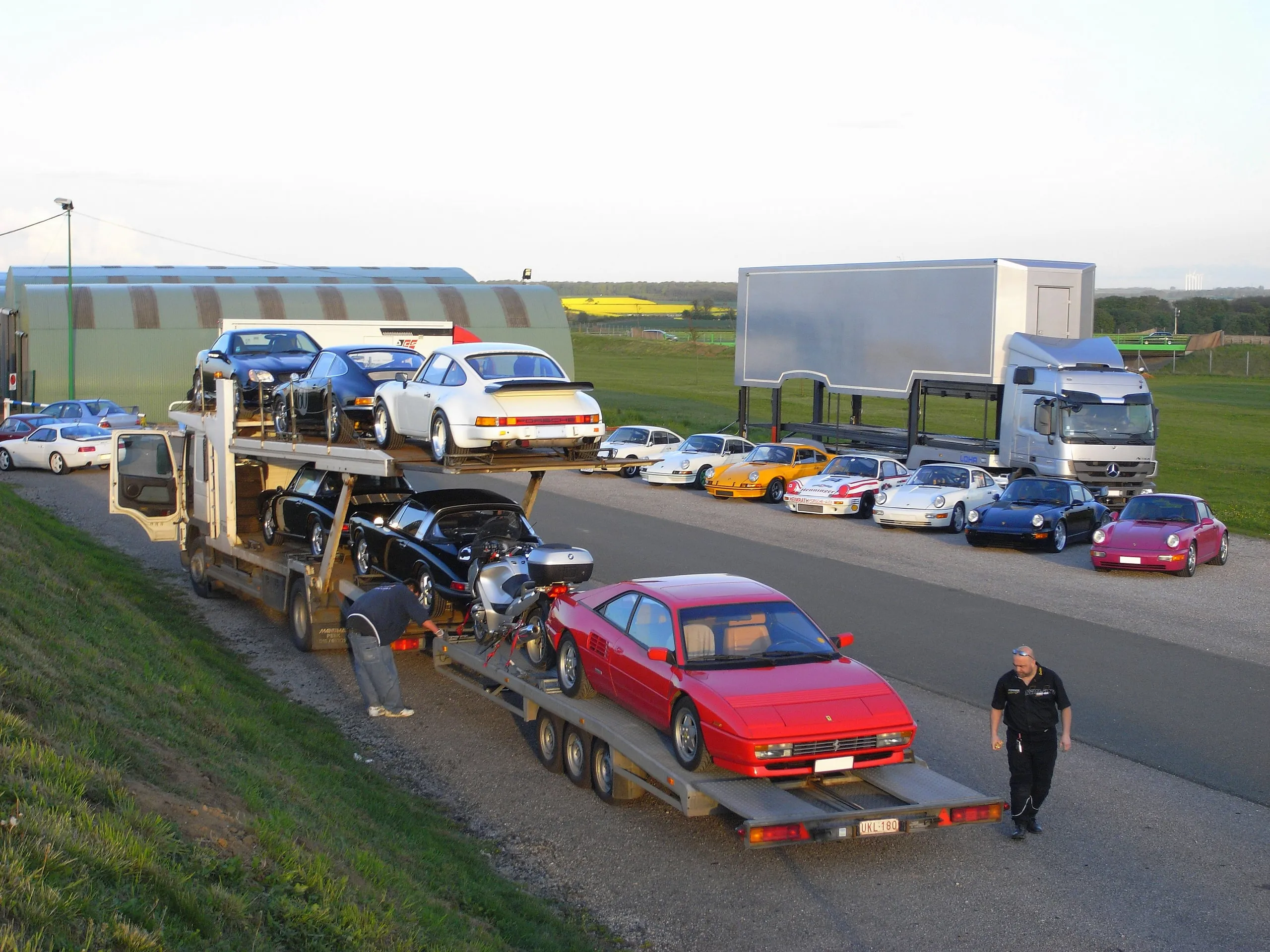 Camion chargé au bord d'une route, une file de voitures de sport attend encore