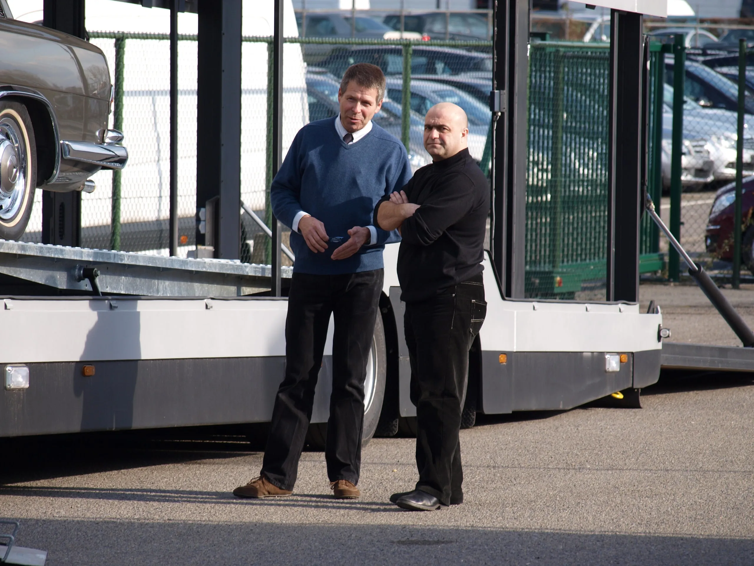 Twee mannen schudden elkaar de hand voor een open transporter met een klassieke wagen