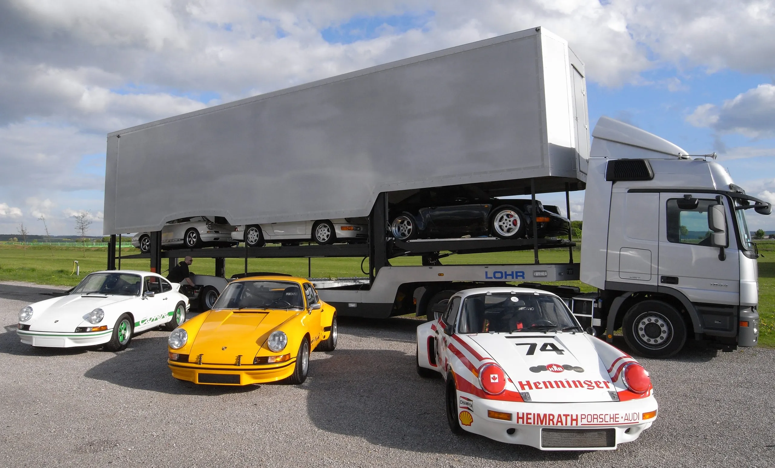 Trois Porsche 911 alignées devant un camion chargé sous un ciel gris