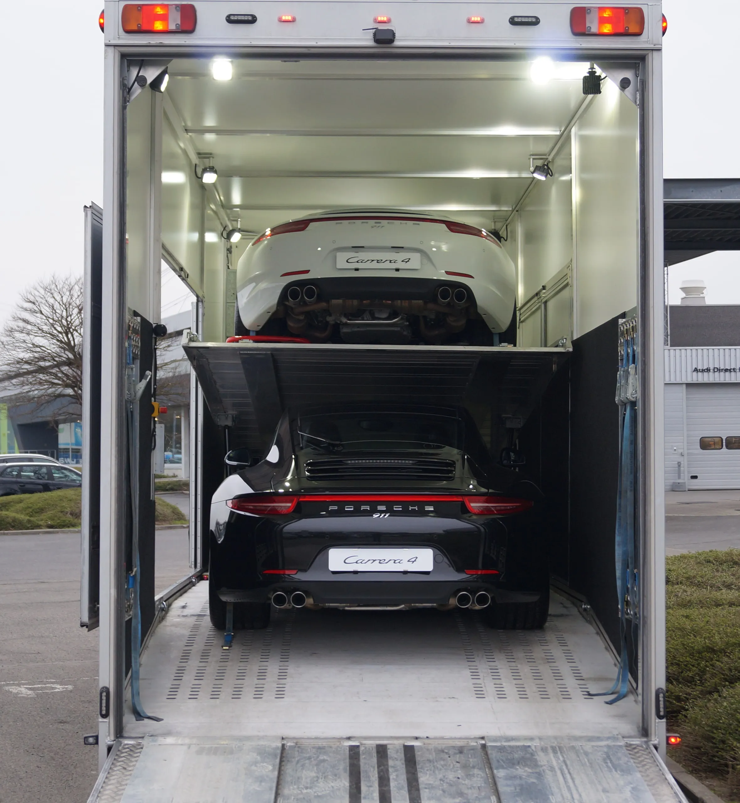 Twee Porsches op de twee dekken van een verlichte gesloten transporter, 's nachts