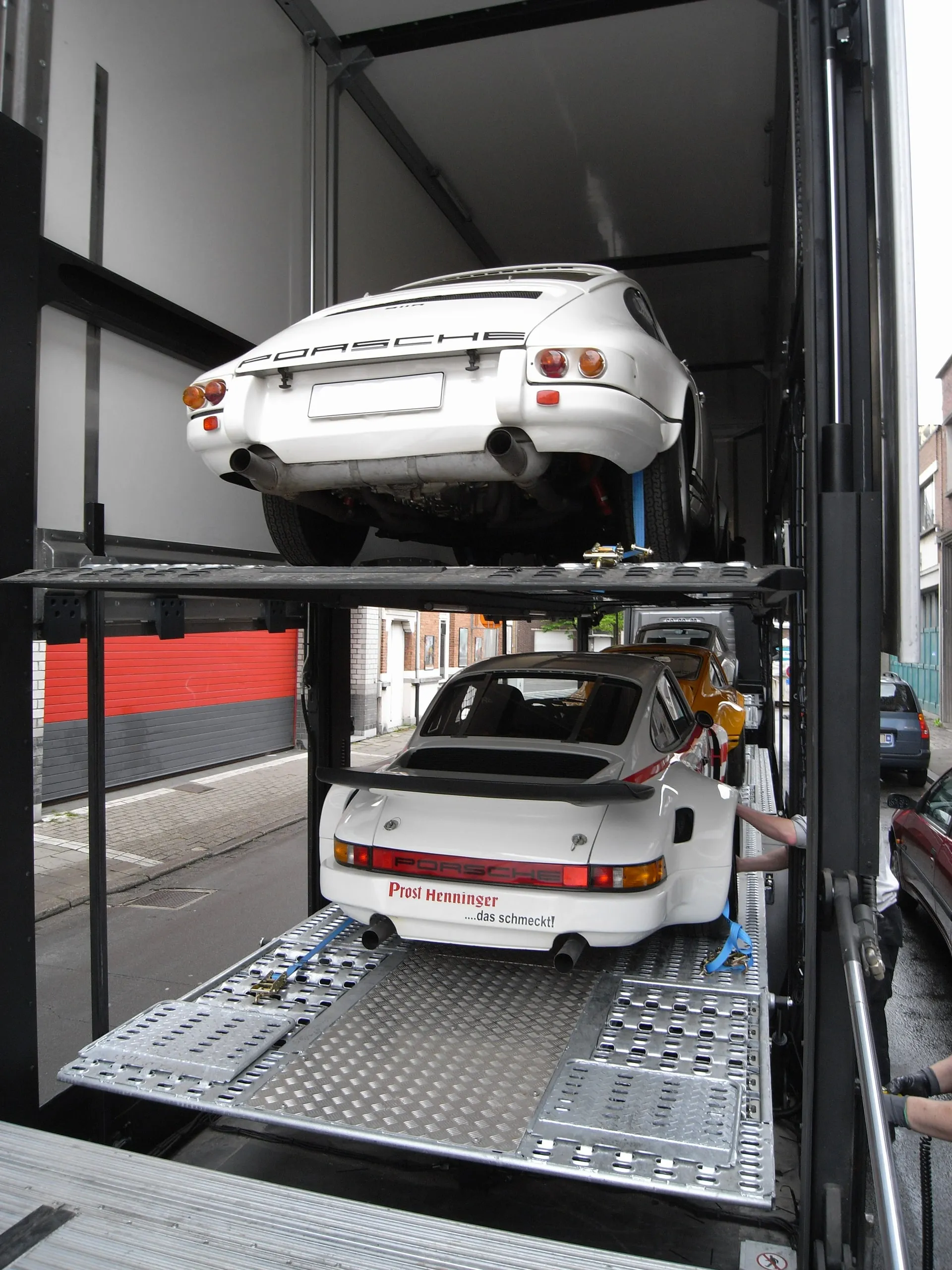 Twee witte Porsche 911's op de twee dekken van een gesloten transporter, platen neer